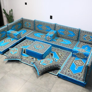May include: A modular seating set with a vibrant blue and patterned design. The set includes multiple cushions, floor cushions, and a central square ottoman. The cushions and floor coverings feature a geometric pattern in black, white, and gold. The set is arranged on a gray tiled floor.