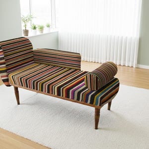 Puede incluir: Una chaise longue decorativa con un patrón de rayas multicolores. El mueble tiene un marco y patas de madera, con un reposabrazos enrollado y un cojín. Se encuentra sobre una alfombra blanca en una habitación con ventana.