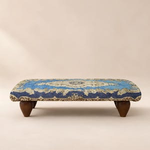 May include: A rectangular low-profile footstool featuring a vibrant blue and beige patterned fabric upholstery with an intricate floral design. The piece is supported by four short, rounded, dark brown wooden legs that provide a stable base.