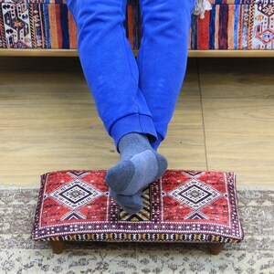 May include: A rectangular footstool with a red and white geometric pattern. The footstool has a dark burgundy border and is supported by four small wooden legs. The footstool is placed on a patterned rug.