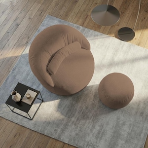 Pu&ograve; includere: Una moderna poltrona in velluto a coste marrone con un pouf rotondo abbinato. La sedia ha uno schienale curvo e un piccolo cuscino attaccato. Il set &egrave; posto su un tappeto grigio chiaro, con un piccolo tavolino nero e pavimento in legno sullo sfondo.