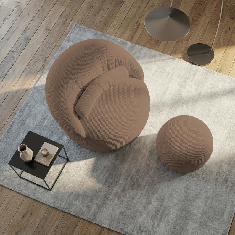 Pu&ograve; includere: Una moderna poltrona in velluto a coste marrone con un pouf rotondo abbinato. La sedia ha uno schienale curvo e un piccolo cuscino attaccato. Il set &egrave; posto su un tappeto grigio chiaro, con un piccolo tavolino nero e pavimento in legno sullo sfondo.