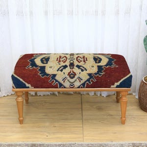 Könnte beinhalten: Ein rechteckiger Hocker mit gemusterter Oberseite in Rot-, Blau- und Beigetönen. Der Hocker hat einen Holzrahmen und vier kurze, hellfarbige Beine. Das Design auf der Oberseite scheint ein traditionelles Teppichmuster zu sein.
