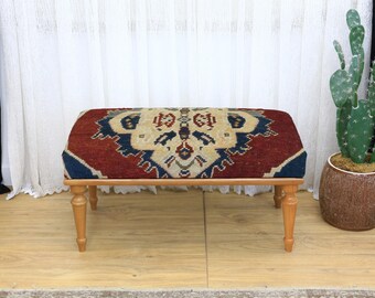 Puede incluir: Un otomán rectangular con una parte superior estampada en tonos de rojo, azul y beige. El otomán tiene un marco de madera y cuatro patas cortas de color claro. El diseño en la parte superior parece ser un patrón de alfombra tradicional.