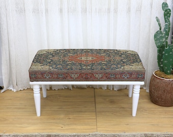 Può includere: Un pouf rettangolare con piano fantasia e gambe bianche. Il piano presenta un design verde scuro, rosso e oro che ricorda un tappeto vintage. Il pouf è un elemento d'arredo decorativo.