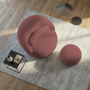 May include: A modern, round armchair and matching ottoman in a dusty rose color. The chair has a curved back and a small, attached pillow. The ottoman is a simple, solid sphere. Both are on a light gray rug.