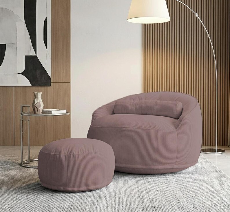 May include: A mauve corduroy beanbag chair with a matching cylindrical pillow and a round ottoman. The chair and ottoman are on a light gray rug. A modern floor lamp and side table are in the background.