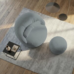 Pu&ograve; includere: Una moderna poltrona imbottita grigio chiaro con un pouf abbinato. La poltrona ha una forma arrotondata con poggiatesta e bracciolo integrati. Il pouf &egrave; un piccolo poggiapiedi rotondo. Un piccolo tavolino nero con oggetti decorativi &egrave; nelle vicinanze.