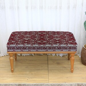May include: A rectangular ottoman with a patterned, burgundy fabric top and light brown wooden legs. The fabric features a detailed, repeating design in shades of burgundy, cream, and grey. The ottoman is a decorative furniture piece.