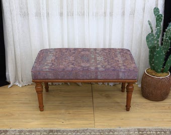 May include: A rectangular ottoman with a patterned, purple upholstered top and four turned wooden legs. The ottoman is a low, decorative piece of furniture, suitable for a living room or bedroom. The legs are a warm brown color.