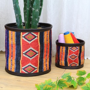Op de afbeelding: Twee cilindrische opbergmanden van stof met een kleurrijk geometrisch patroon. De grotere mand is gevuld met een cactusplant in een pot. De kleinere mand is gevuld met kleurrijk garen.