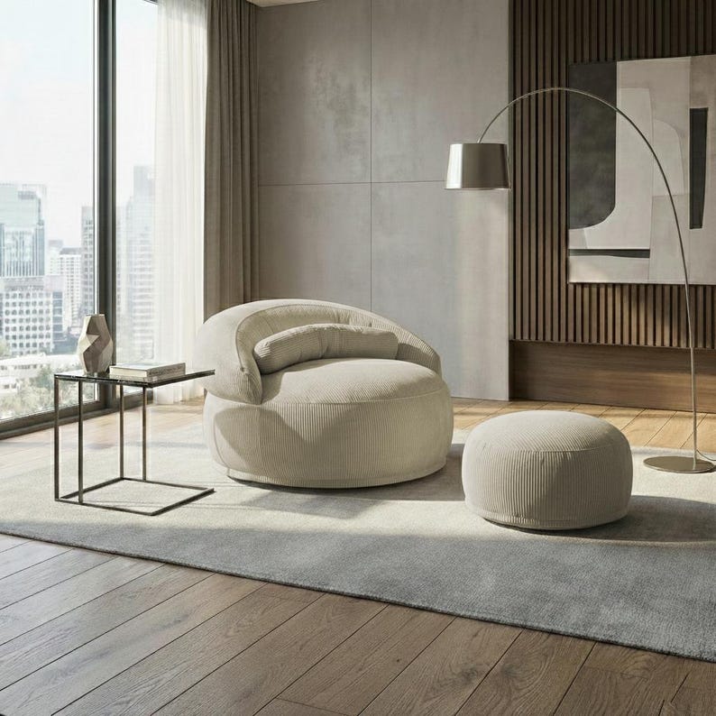 May include: A modern living room scene featuring a beige corduroy armchair with a matching ottoman. A small black side table with a glass top and a silver arc floor lamp are also present. The room has a neutral color palette with a city view.