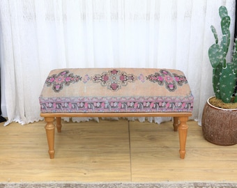 May include: A decorative bench with a patterned top and wooden legs. The bench features a beige background with pink and purple floral designs. The legs are a light brown color. A cactus in a brown pot is in the background.