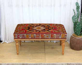 Puede incluir: Un otomán rectangular con estructura de madera y cuatro patas. La parte superior está cubierta con una tela estampada vibrante con diseños geométricos rojos, naranjas, amarillos y negros. Un cactus en maceta está cerca.