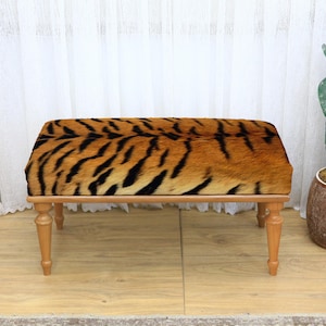 May include: A rectangular bench with a tiger print seat cushion and light brown wooden legs. The bench is set against a backdrop of white curtains and a potted cactus.
