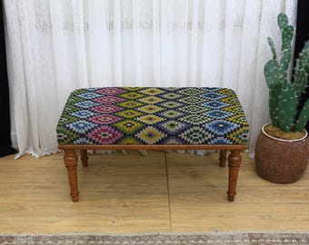 May include: A rectangular upholstered ottoman with a colorful geometric pattern in shades of blue, green, yellow, and pink. The wooden frame has four turned legs. The ottoman is approximately 30 inches long.