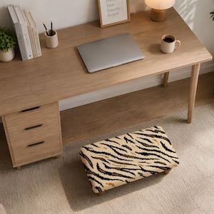 May include: A rectangular footrest featuring a beige and black tiger stripe pattern, positioned on a light-colored rug in front of a wooden desk. The footrest has a cushioned top surface and is supported by four short, sturdy wooden legs.