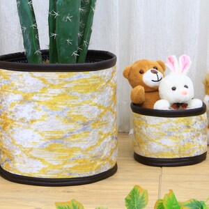 May include: Two yellow and white patterned fabric storage baskets with brown trim. One basket is filled with a brown teddy bear and a white bunny rabbit. The other basket is empty and sits next to a cactus plant.
