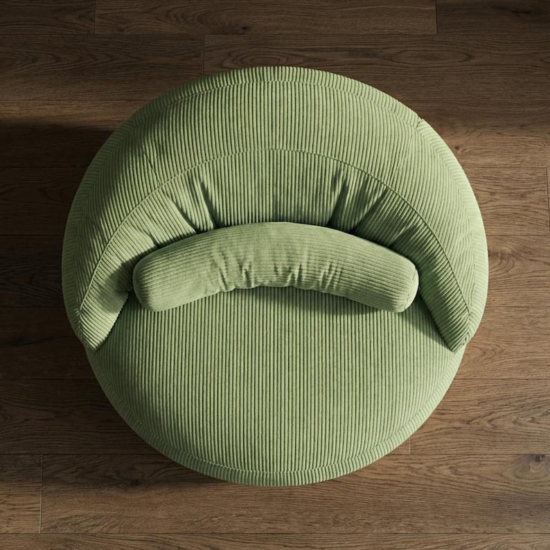 May include: A round, light green corduroy chair with a curved backrest and a cylindrical bolster pillow. The chair is set against a dark wood floor, offering a comfortable and stylish seating option for any living space.