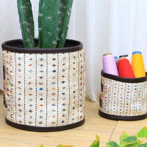 Könnte beinhalten: Zwei gewebte Aufbewahrungskörbe mit geometrischem Muster. Der größere Korb ist mit einer Kaktuspflanze gefüllt, der kleinere Korb mit buntem Garn.