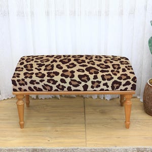 May include: A rectangular ottoman with a leopard print fabric top and light brown wooden legs. The fabric features a pattern of brown spots on a tan background. The ottoman is a decorative furniture piece.