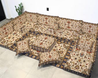 Può includere: Un set di sedute modulari a pavimento con un motivo floreale beige, rosso e blu. Il set comprende più cuscini e una grande base simile a un tappeto, creando un'area salotto confortevole ed elegante. Il design ricorda l'arredamento tradizionale mediorientale.