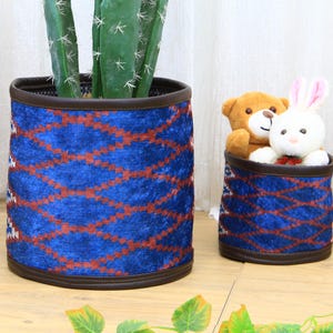 Könnte beinhalten: Zwei blaue Stoff-Pflanztöpfe mit einem geometrischen Muster in Rot und Braun. Der größere Topf enthält einen Kaktus, und der kleinere Topf enthält Stofftiere. Die Töpfe haben eine dunkelbraune Umrandung.