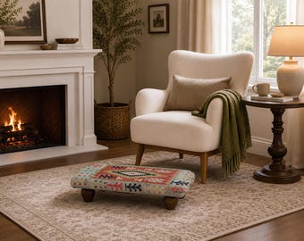 May include: A low rectangular footstool featuring a multi-colored woven fabric top with geometric patterns in red, blue, and green, supported by four short, dark brown wooden feet. The stool is positioned in front of a cream-colored armchair.