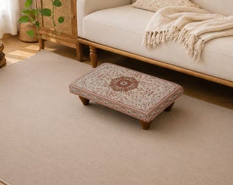 May include: A rectangular footstool featuring a woven fabric top with a traditional intricate floral pattern in shades of red, blue, and cream. The low-profile stool is supported by four short, dark brown wooden legs, providing a stable base.