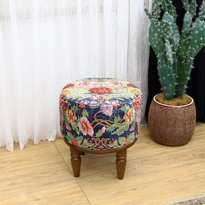 Peut inclure: Un pouf rond à motifs floraux avec des pieds en bois. Le pouf a un fond bleu foncé avec des fleurs colorées et un motif floral rouge et rose au centre.