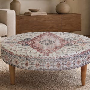 May include: A round ottoman featuring a cream-colored fabric top with a faded, intricate medallion pattern in shades of dusty rose, muted blue, and charcoal gray. The piece is supported by four short, light-toned wooden legs.