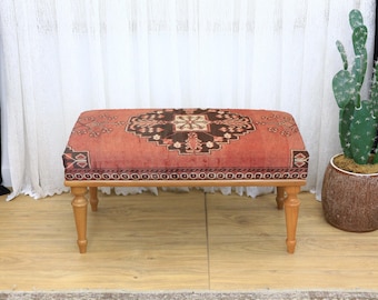 May include: A rectangular ottoman with a patterned, reddish-orange top and light brown wooden legs. The top features a central medallion design in brown and black. The ottoman is placed in front of a white curtain and next to a potted cactus.