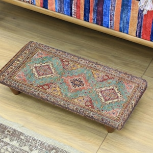 May include: Rectangular, low-profile footstool with a detailed, patterned rug top. The rug features a teal, red, and gold design with a central medallion and corner accents. The stool has short, wooden legs and is set on a wooden floor.