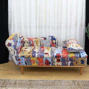 May include: A small, colorful sofa with a patchwork design. The sofa has wooden legs and a matching pillow. The fabric features a mix of patterns and colors, including red, blue, and yellow. A potted cactus sits to the right.