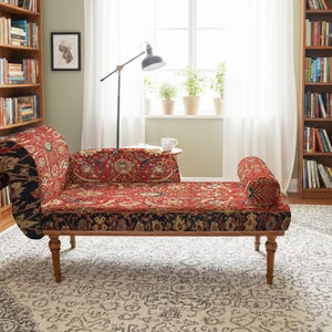 May include: A chaise lounge with a red and black patterned fabric. The wooden frame has curved legs and a rolled arm. The room features bookshelves, a patterned rug, and a window with plants.