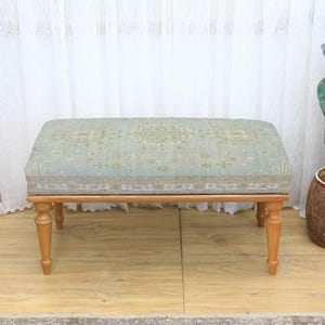 May include: A rectangular ottoman with a light blue and beige patterned top and four wooden legs. The ottoman is placed in front of a white curtain and a cactus in a brown pot.