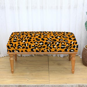 May include: A rectangular bench with a leopard print seat cushion in shades of orange, black, and white. The bench has four wooden legs and is placed in front of a white curtain. A cactus in a brown pot is to the right.