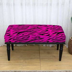 May include: A black-legged bench with a vibrant pink seat featuring a bold black zebra stripe pattern. The bench is a decorative furniture piece, suitable for various interior settings. A cactus in a brown pot is in the background.
