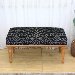 May include: A rectangular ottoman with a black and white patterned top and light brown wooden legs. The fabric design features a repeating geometric pattern. The ottoman is a decorative furniture piece.