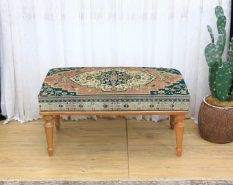 May include: A rectangular ottoman with a patterned top in shades of teal, peach, and cream. The ottoman has four light-colored wooden legs. The design on the top resembles a traditional rug pattern. A cactus in a brown pot is in the background.