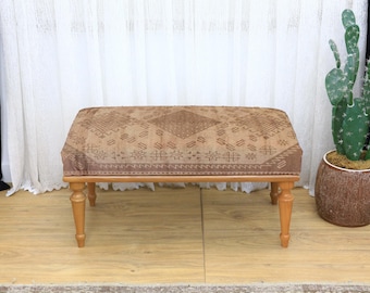 Puede incluir: Un otomán rectangular con una parte superior estampada y patas de madera. La parte superior presenta un diseño geométrico marrón y beige. Las patas son de color marrón claro y ligeramente cónicas. El otomán está colocado frente a una cortina blanca.
