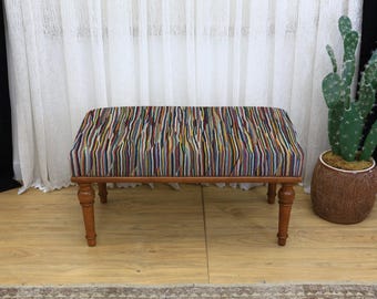 May include: A rectangular bench with a multi-colored striped fabric seat and a wooden frame. The bench has four turned legs and is placed on a light-colored wooden floor. A cactus in a brown pot is to the right.