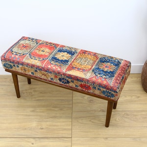 May include: A wooden bench with a colorful patterned cushion. The cushion has a geometric design with red, blue, and yellow colors. The bench is sitting on a wooden floor.