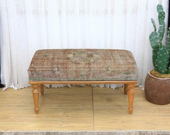 Puede incluir: Un otomán rectangular con una parte superior de tela desgastada y estampada en tonos marrones, verdes y beige. El otomán tiene un marco de madera y cuatro patas torneadas. Un cactus en una maceta marrón está a la derecha.