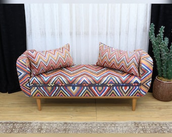 May include: A small, upholstered bench with a colorful zigzag pattern in shades of orange, red, blue, and yellow. The bench has two matching throw pillows and wooden legs. The bench is approximately 48 inches long.