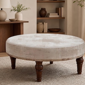 May include: A round ottoman featuring a textured, light beige fabric top with a subtle distressed pattern. The piece is supported by four dark brown wooden legs with a turned, spindle-style design, providing a stable base for this furniture item.