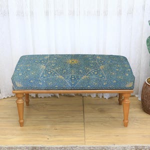 May include: A rectangular ottoman with a blue patterned top and light brown wooden legs. The blue fabric features a gold starburst design. The ottoman is a decorative furniture piece.