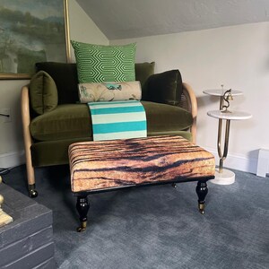 May include: A green velvet sofa with a tiger-print ottoman. The sofa has a curved wooden frame and is adorned with green and patterned pillows and a turquoise and white striped throw. A small side table is visible.