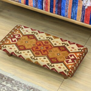 Peut inclure: Un repose-pieds rectangulaire avec un dessus en tissu à motifs beiges, oranges et rouges. Le repose-pieds a un cadre en bois et des pieds courts et arrondis. Le motif présente des formes géométriques et une bordure.