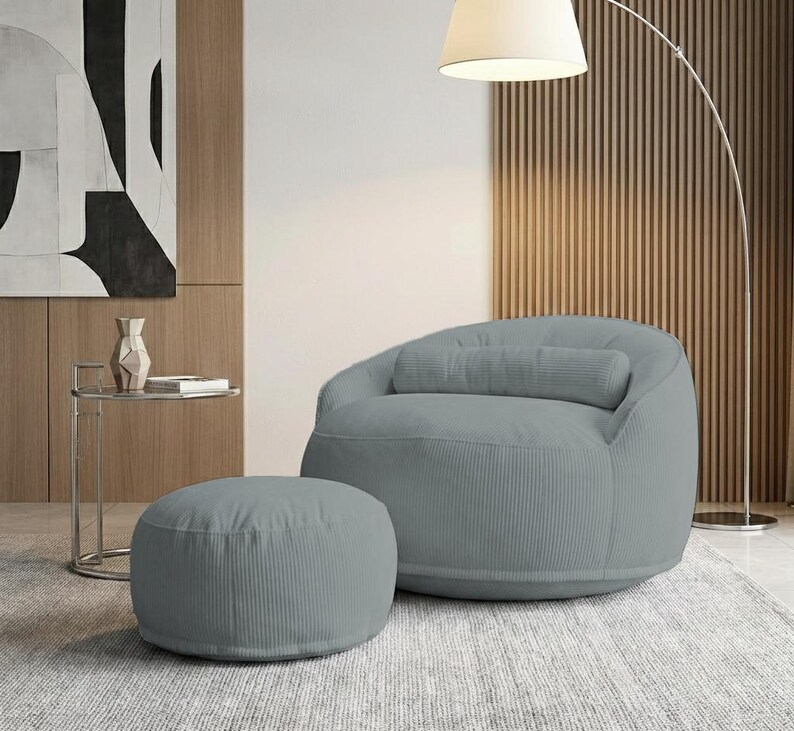 Pode incluir: Uma poltrona beanbag de veludo cotel&ecirc; cinza claro com um pufe a condizer. A cadeira tem encosto arredondado e uma almofada cil&iacute;ndrica. O pufe &eacute; circular. Uma moderna lumin&aacute;ria de ch&atilde;o e uma mesa lateral est&atilde;o no fundo.